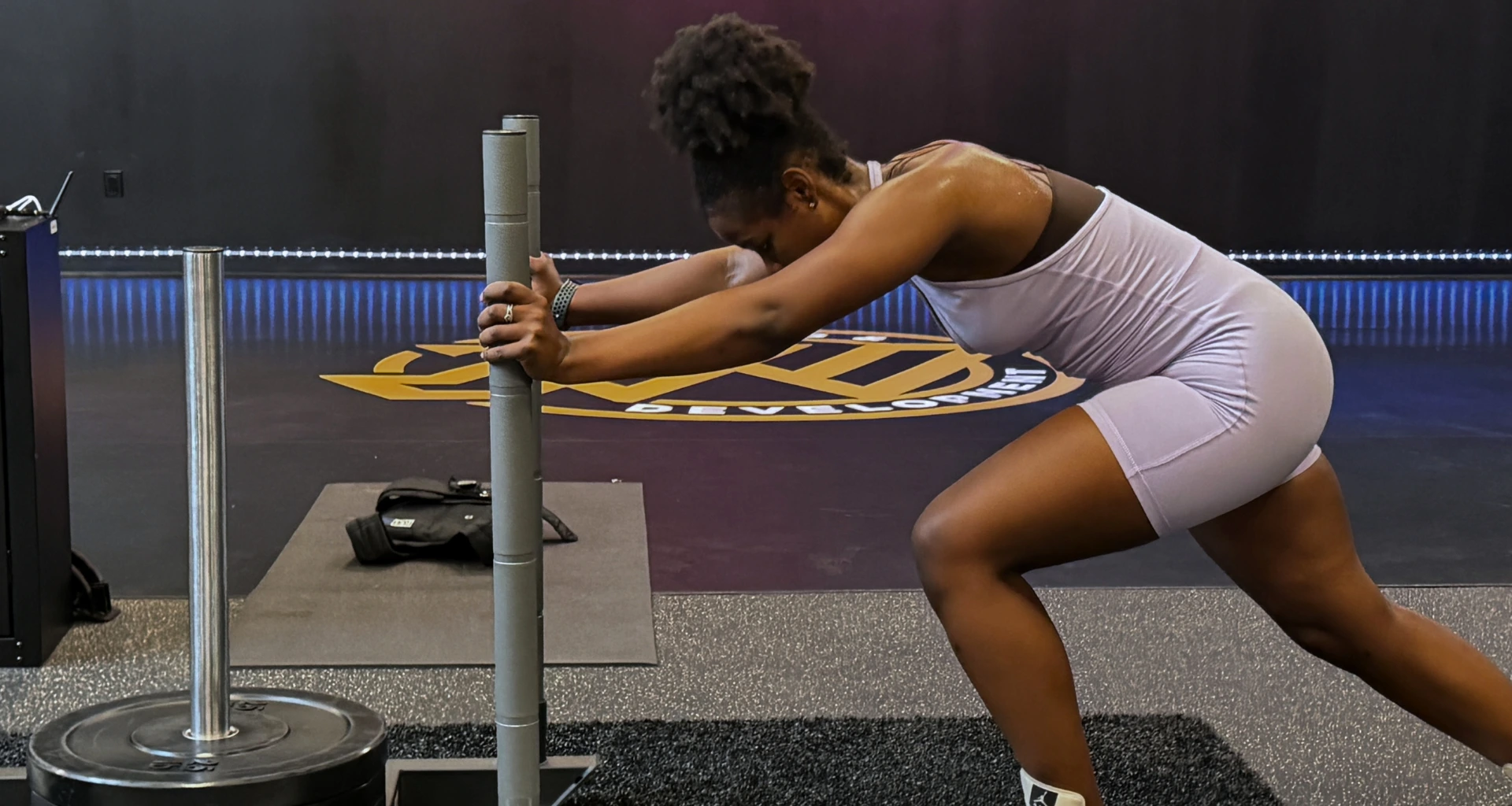 A woman in athletic wear pushes a weighted sled in a gym, leaning forward with both hands on the sled handles, demonstrating strength and focus during her workout—embodying the dedication and power of a true JW H.E.R.O.