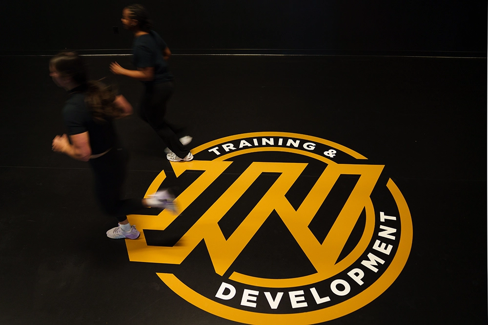 Two people run on a black floor featuring a large yellow logo with the words JW Training and Development in a circle around a stylized design, capturing the energetic atmosphere highlighted on the JW About Page.