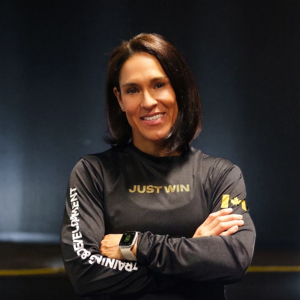 A woman with straight brown hair stands smiling with arms crossed, wearing a black long-sleeve shirt that says JUST WIN and an Apple Watch. JW Training and Development