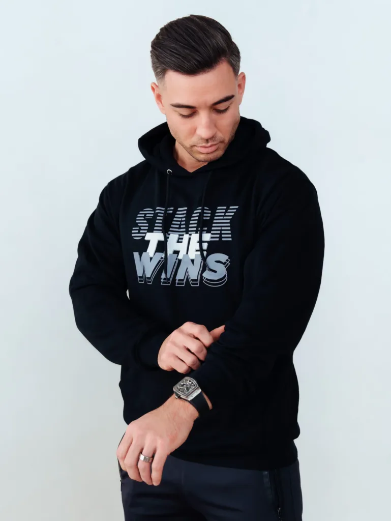 Man in a black hoodie with STACK THE WINS text looks down while adjusting his sleeve. He wears a watch and has short, neatly styled hair. Plain, light background. JW Training and Development