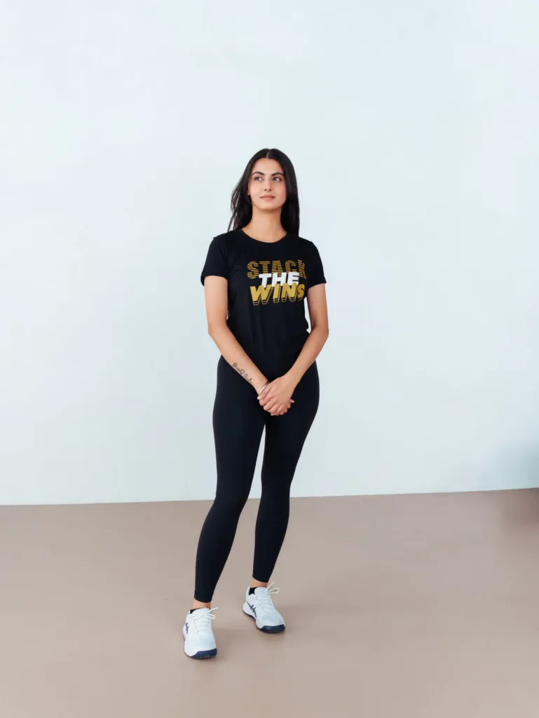 A woman stands indoors against a plain background, wearing a black Stack The Wins T-shirt, black leggings, and white sneakers. Her hands are clasped in front of her. JW Training and Development