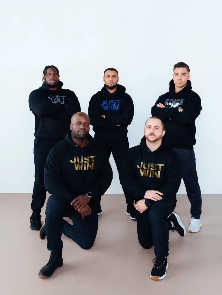 Five men in black hoodies pose together, with two kneeling in front and three standing behind. The hoodies have JUST WIN or similar motivational phrases printed on them. They are indoors, against a plain white wall. JW Training and Development