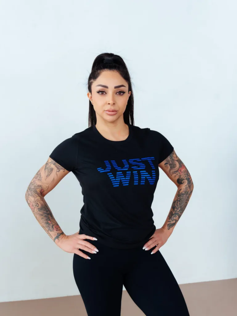 A woman with long dark hair in a ponytail stands confidently with hands on hips, wearing a black JUST WIN t-shirt and black pants. She has visible tattoos on both arms and is standing against a light background. JW Training and Development