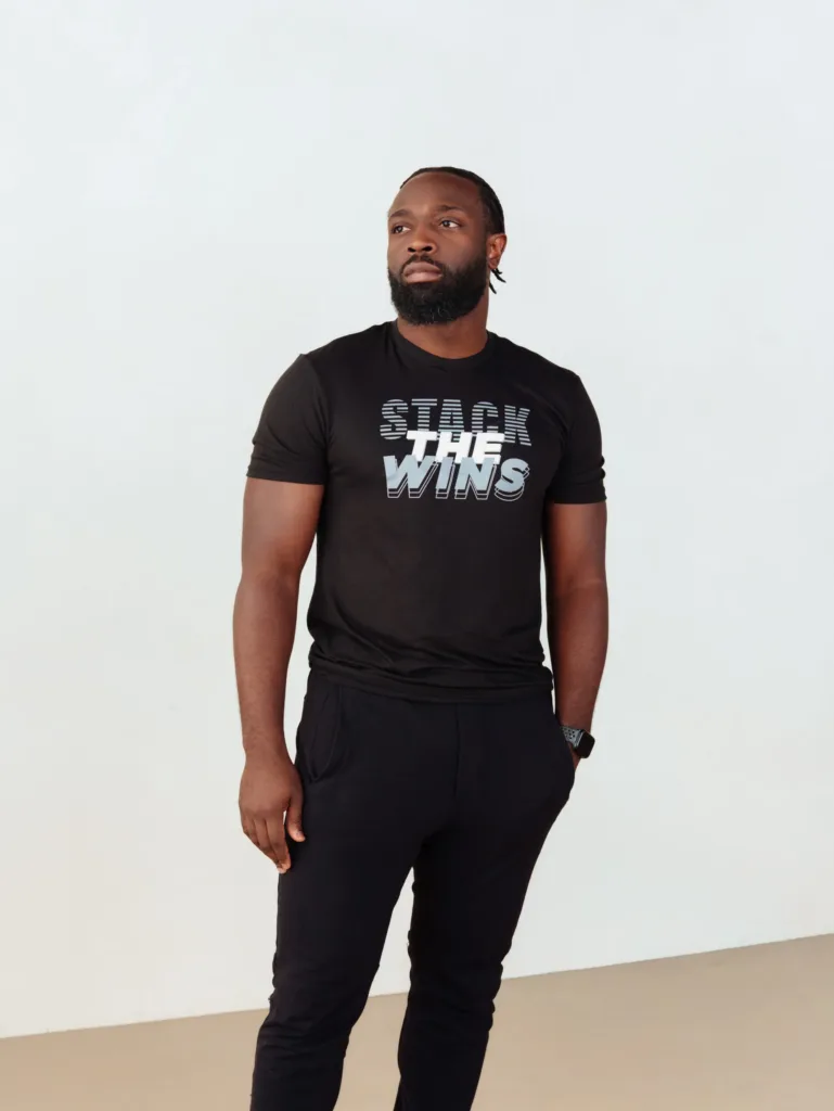 A man stands against a plain background wearing a black T-shirt that says STACK THE WINS in bold white and gray letters. He has a neutral expression and his left hand is resting by his side. JW Training and Development