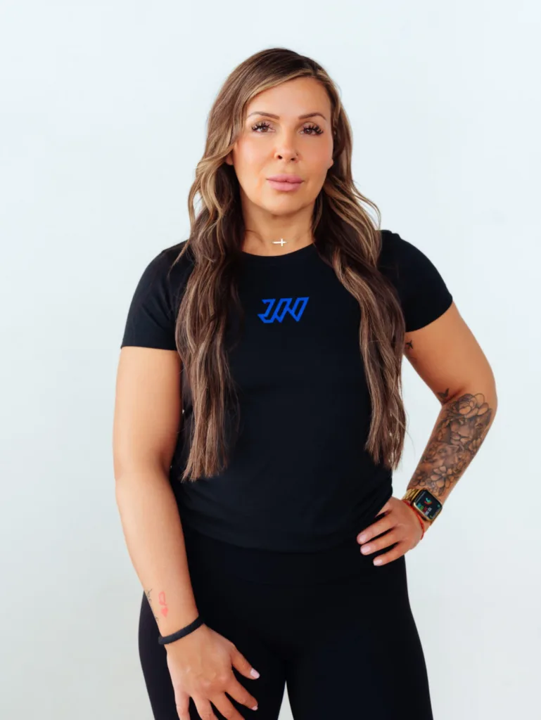 A woman with long brown hair wearing a black t-shirt and leggings stands against a white background. She has tattoos on her forearm and is wearing a smartwatch and a cross necklace. JW Training and Development