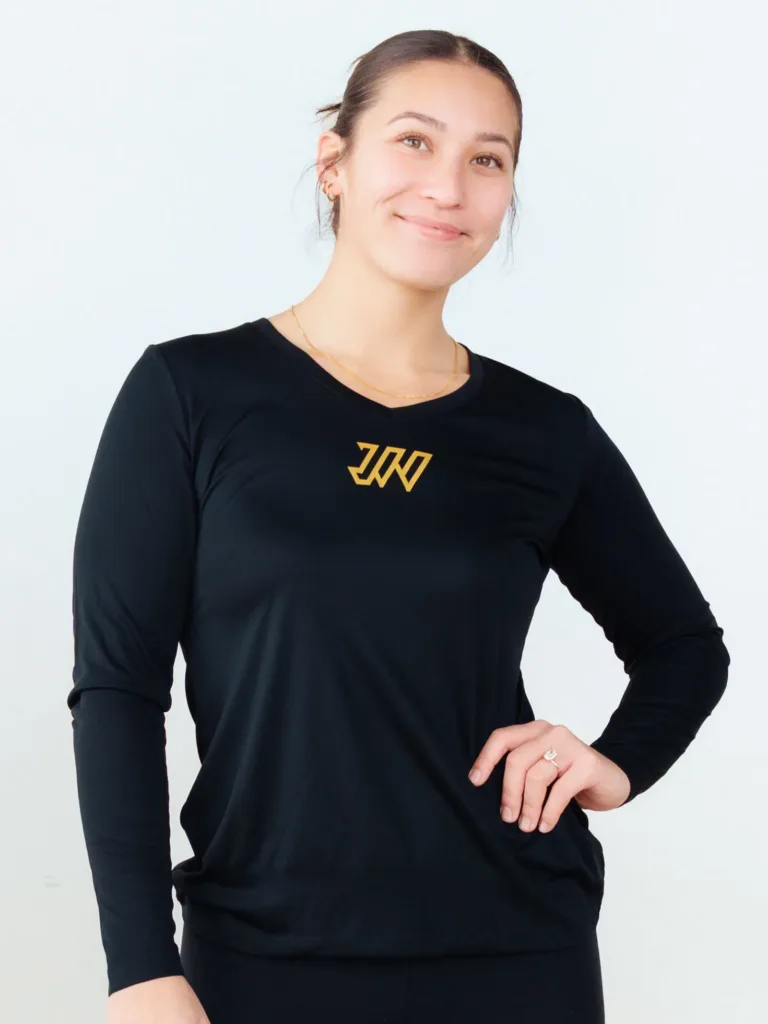 A person in a black long-sleeve shirt with a gold geometric logo stands against a plain light background, smiling with one hand on their hip. JW Training and Development