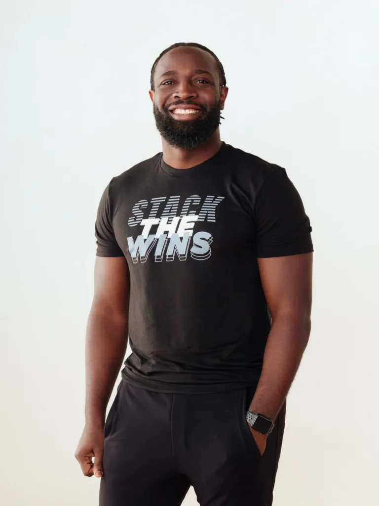 Smiling man with a beard stands against a plain background, wearing a black Stack The Wins t-shirt, black pants, and a smartwatch. JW Training and Development