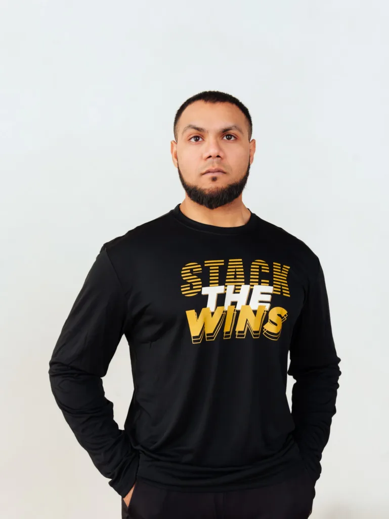 A person stands facing forward, wearing a black long-sleeve shirt with the words STACK THE WINS in bold yellow and white text. The background is plain and light-colored. JW Training and Development