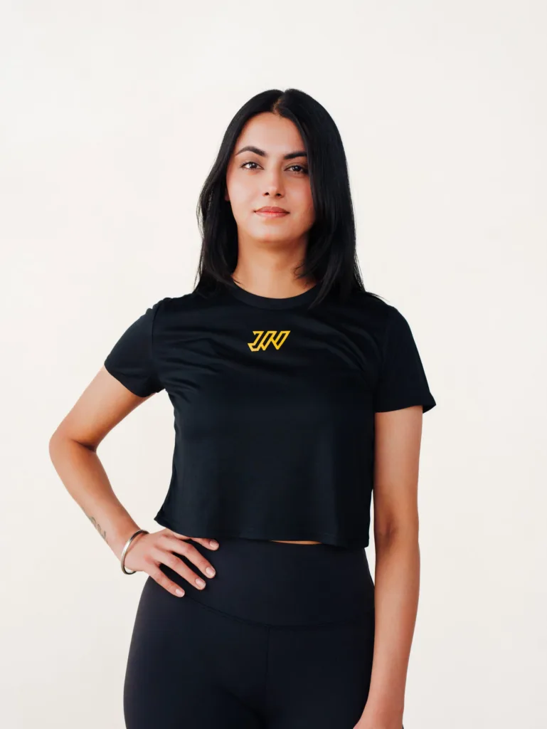 Woman with straight black hair wearing a black crop top and leggings stands confidently against a plain white background, with one hand on her hip. JW Training and Development
