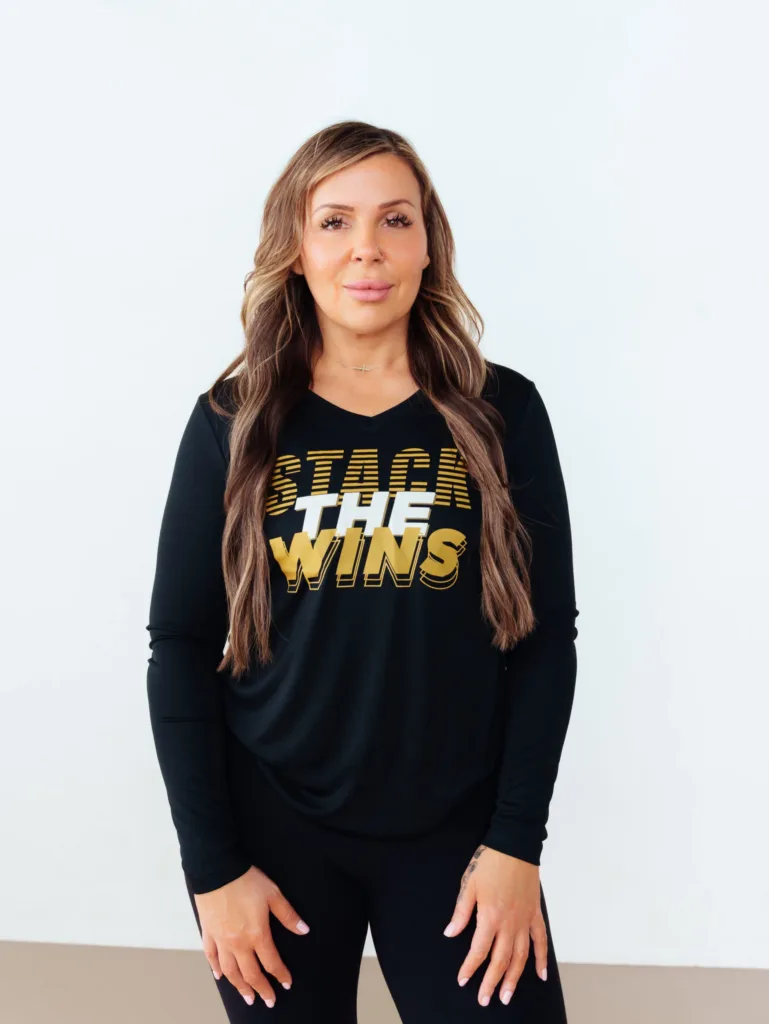 A woman with long wavy hair wearing a black long-sleeve shirt that says STACK THE WINS in gold and white letters stands against a plain light-colored background. JW Training and Development