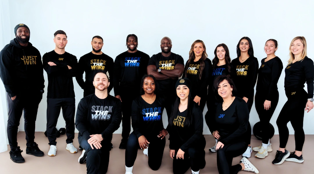 Thirteen people in black athletic wear pose together, smiling. Some shirts read Steal the Wins or Just Win. Six people are standing, and seven are kneeling or sitting in front, all against a plain light background. JW Training and Development